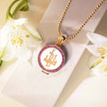 Gold necklace with a circular pendant featuring initials, surrounded by white flowers on a light background