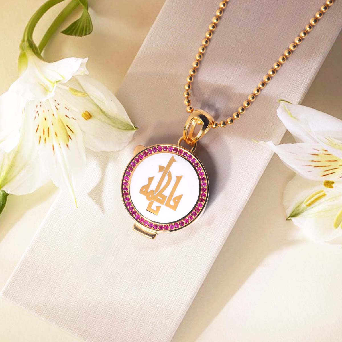 Gold necklace with a circular pendant featuring initials, surrounded by white flowers on a light background